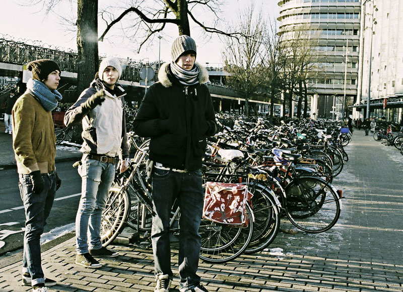 Three young adults standing in front of a large bicycle parking area, dressed in winter clothing. Bicycles are lined up behind them, and there are trees and buildings in the background.